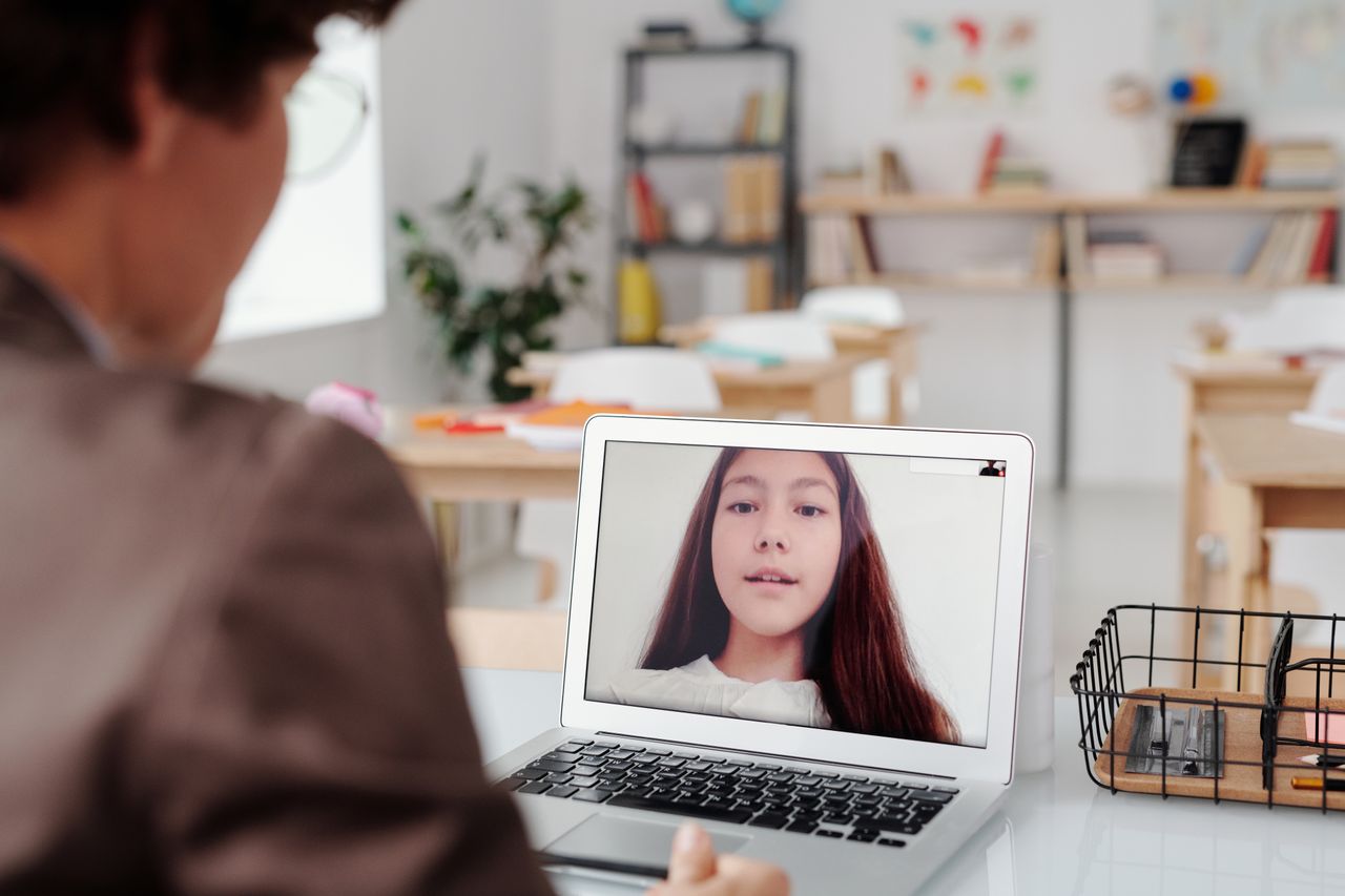 Services Teacher using laptop for virtual lesson with female student on screen in classroom setting.